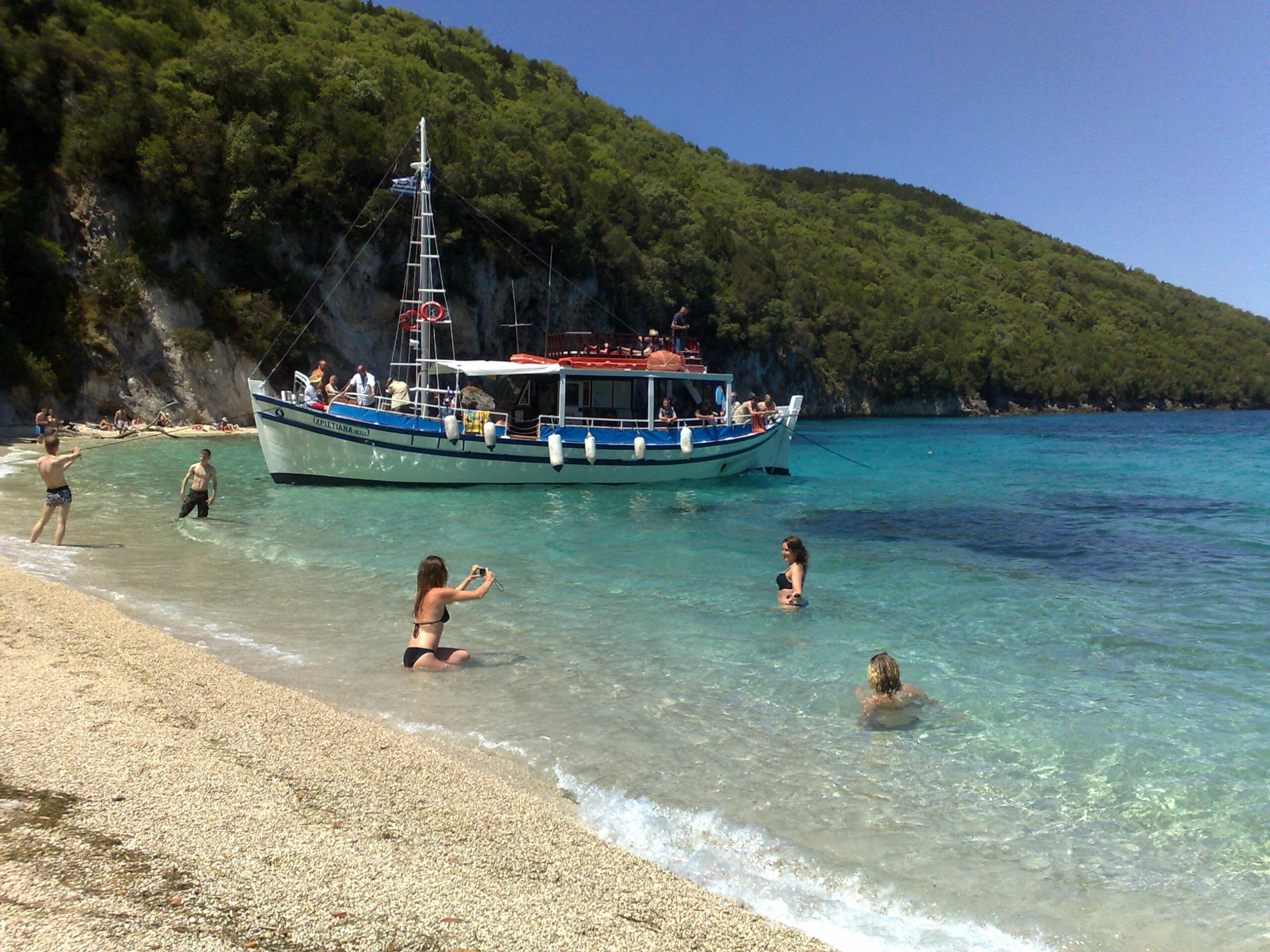 Tour boat anchored at the South Lagoon Sandbank in Corfu with crystal-clear shallow waters and passengers swimming during a half-day boat excursion.